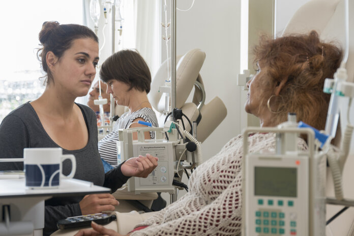 Eine ältere Frau liegt auf einer Liege und erhält eine Chemotherapie, während eine junge Frau bei ihr sitzt.