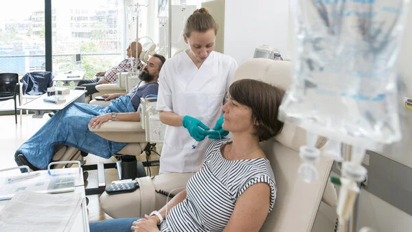 Eine Patientin sitzt in einer Chemo-Ambulanz mit anderen Patienten. Eine Krankenschwester bereitet ihre Infusion vor.