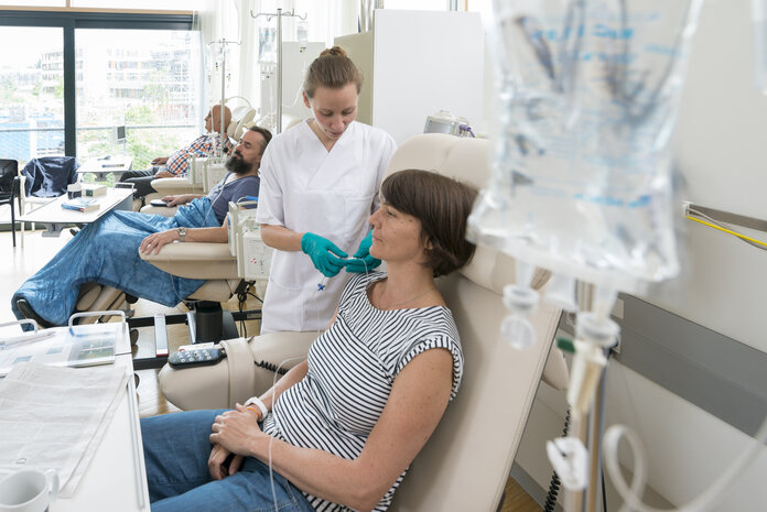Eine Patientin sitzt in einer Chemo-Ambulanz mit anderen Patienten. Eine Krankenschwester bereitet ihre Infusion vor.