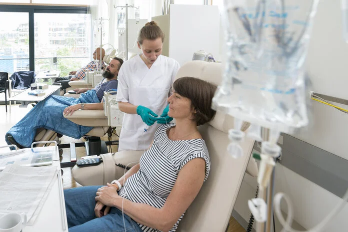 Eine Patientin sitzt in einer Chemo-Ambulanz mit anderen Patienten. Eine Krankenschwester bereitet ihre Infusion vor.