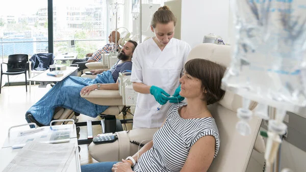 Chemoinfusin vorbereiten, © Krebsinformationsdienst, DKFZ; Foto: Tobias Schwerdt Eine Patientin sitzt in einer Chemo-Ambulanz mit anderen Patienten. Eine Krankenschwester bereitet ihre Infusion vor.