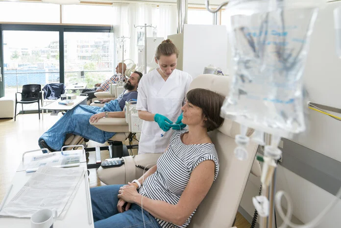 Chemoinfusin vorbereiten, © Krebsinformationsdienst, DKFZ; Foto: Tobias Schwerdt Eine Patientin sitzt in einer Chemo-Ambulanz mit anderen Patienten. Eine Krankenschwester bereitet ihre Infusion vor.