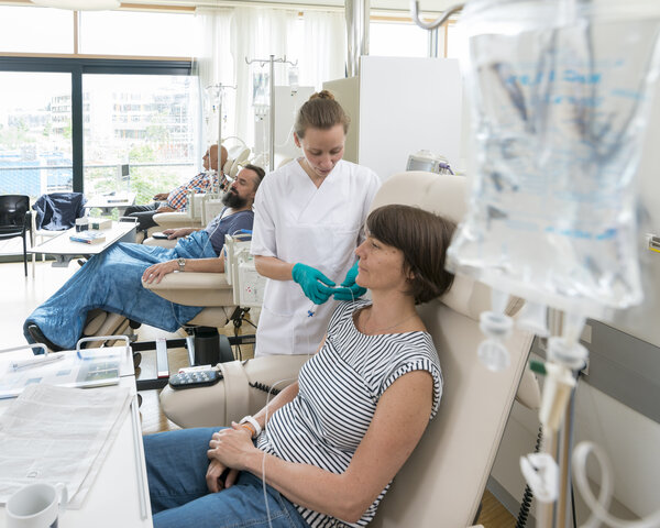 Eine Patientin sitzt in einer Chemo-Ambulanz mit anderen Patienten. Eine Krankenschwester bereitet ihre Infusion vor.