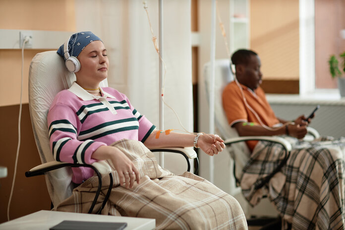 Eine Frau sitzt mit Kopfhörern bei der Chemotherapie, im Hintergrund schaut ein weiterer Patient auf sein Handy.