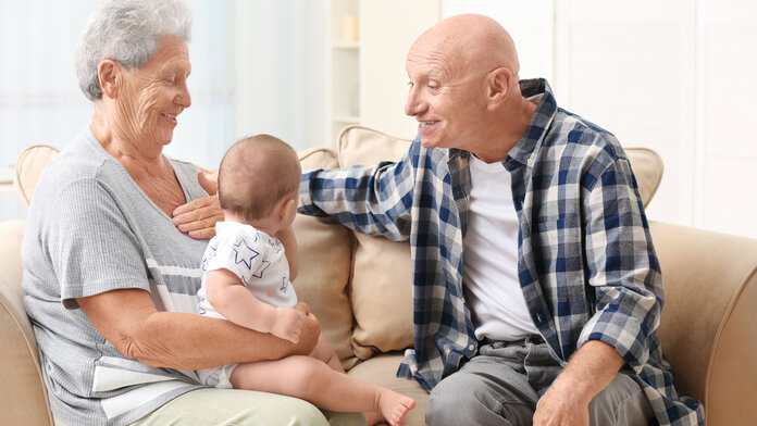 Oma hat Enkel auf dem Schoß sitzen, gegenüber sitzt der Opa und lächelt das Baby an.