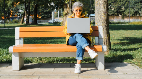 Eine ältere Frau sitzt mit ihrem Laptop auf einer Parkbank.