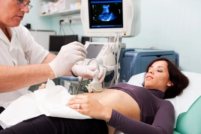 Ultraschall-unterstützte Biopsie, © choja, iStock Eine junge Frau liegt auf einer Untersuchungsliege. Ein Arzt hält einen Ultraschallkopf auf ihren Bauch und entnimmt mit einer langen Nadel eine Probe. Im Hintergrund zeigt ein Bildschirm ein Ultraschallbild.