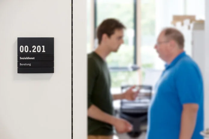Mann im Gespräch mit einem Sozialarbeiter, © Krebsinformationsdienst, DKFZ; Foto: Tobias Schwerdt Tür mit Schild "Sozialdienst". Im Hintergrund spricht ein älterer Mann mit einem jungen Solzialarbeiter.
