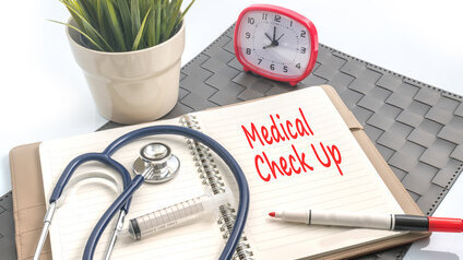 Auf der rechten Seite eines aufgeschlagenen Ringbuchs steht in roter Schrift Medical Check Up, auf der linken Seite liegen ein Stethoskop und eine Spritze.