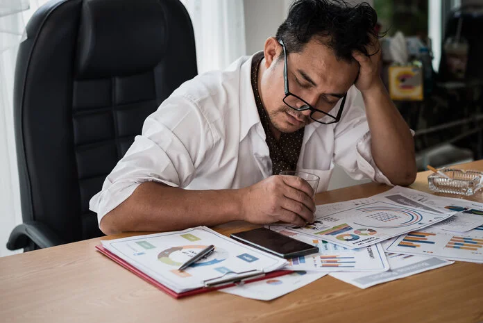 Gestresster Mann, © worawit_j - shutterstock Gestresster Mann am Schreibtisch mit Alkohol und Zigaretten
