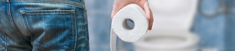 Mann von hinten geht mit einer Rolle Klopapier zur Toilette.