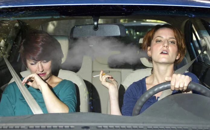 Passivrauchen, © innovatedcaptures, iStock.com Eine junge Raucherin sitz hinter dem Steuer eines Autos - die Beifahrerin wendet sich hustend ab