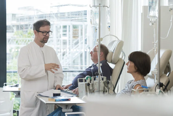 Behandlung in der Tagesklinik , © Krebsinformationsdienst, DKFZ; Foto: Tobias Schwerdt Eine Patientin und ein Patient sitzen nebeneinander in Behandlungsstühlen einer Tagesklinik und erhalten eine Infusion, ein Arzt steht bei der Patientin und erklärt etwas.