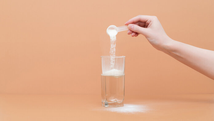 Eine Person schüttet Kollagen-Pulver mit dem Löffel in ein Wasserglas. Auf dem Bild ist nur die Hand der Person zu sehen.
