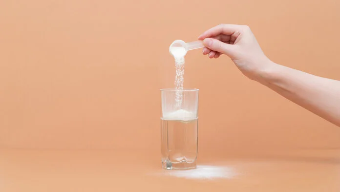 Eine Person schüttet Kollagen-Pulver mit dem Löffel in ein Wasserglas. Auf dem Bild ist nur die Hand der Person zu sehen.