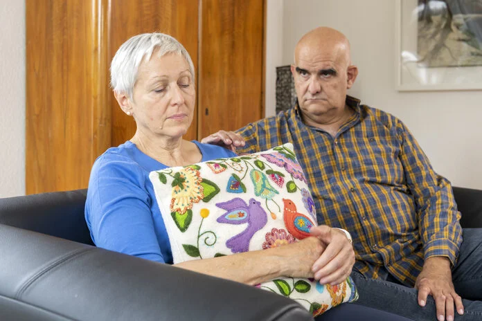 Eine Frau mit erschöpften Blick sitzt auf dem Sofa. Ein Mann legt fürsorglich die Hand auf ihre Schulter.