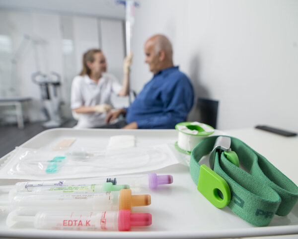 Im Vordergrund ein Tablett mit Blutentnahme-Röhrchen und Besteck zum Blutabhnehmen. Im Hintergrund eine Arzthelferin, die einem Mann Blut abnimmt.