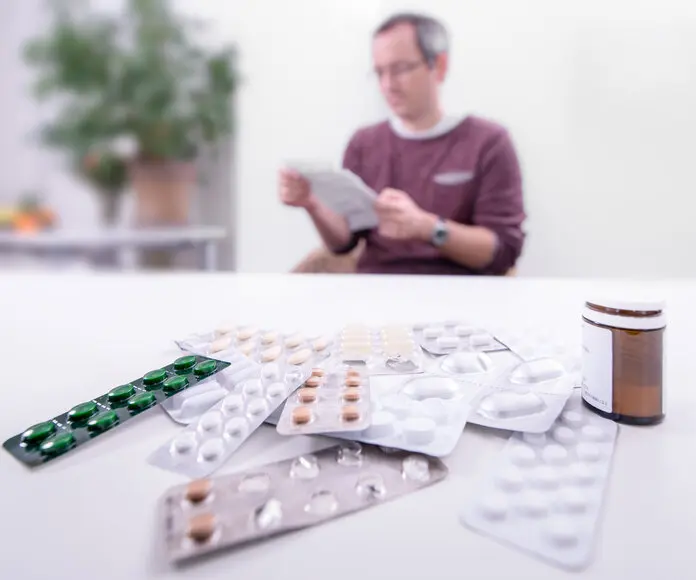 Medikamentenwechselwirkungen, ©Krebsinformationsdienst, DKFZ Auf dem Tisch liegen etliche Tablettenblister, im Hintergrund sitzt ein Patient, der einen Beipackzettel liest.