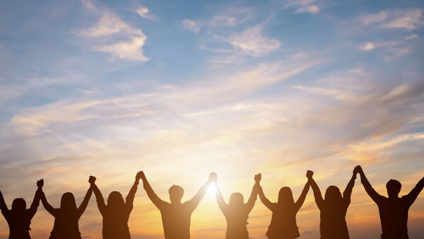 Man sieht die Silhouette einer Menschengruppe vor einem Sonnenuntergang die ihre Arme hoch halten und sich an den Händenfassen.