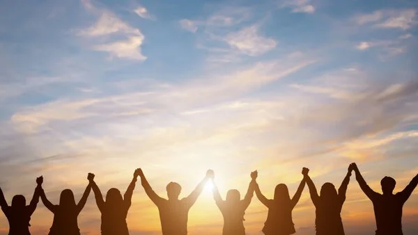 Gemeinschaft, © Guitarfoto studio, Shutterstock Man sieht die Silhouette einer Menschengruppe vor einem Sonnenuntergang die ihre Arme hoch halten und sich an den Händenfassen.