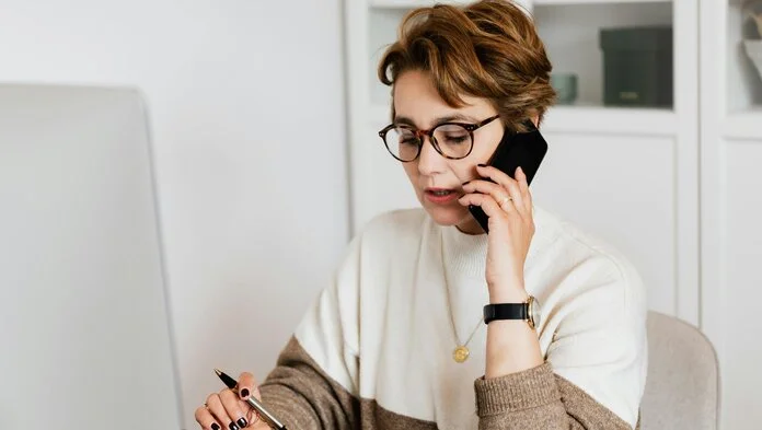 Telefonieren mit Handy, © Karolina Grabowska, Pexels Eine Frau sitzt am Computer und telefoniert mit ihrem Handy am Ohr.