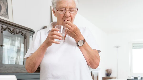 Medikamenten-Einnahme, © Krebsinformationsdienst, DKFZ; Foto: Tobias Schwerdt Ältere Frau nimmt Medikamente mit einem Glas Wasser ein. Vor ihr auf dem Tisch liegt eine Dosierhilfe.