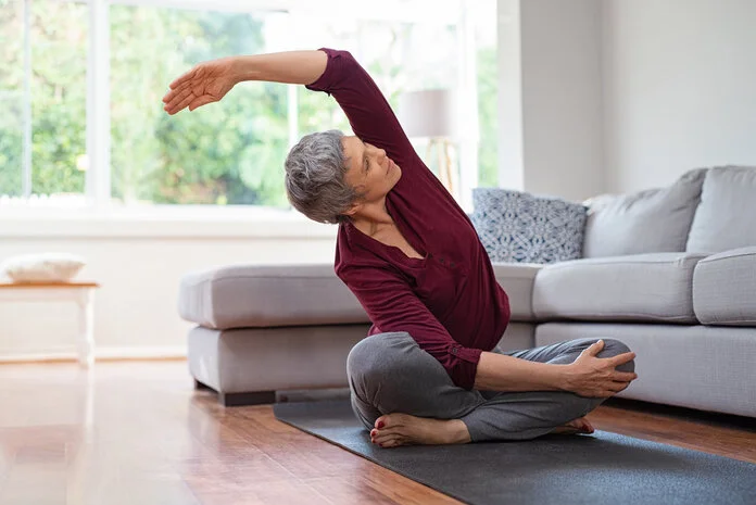 Ältere Frau macht eine Dehnübung auf einer Yogamatte neben einem Sofa.