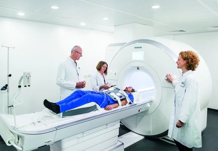 Kernspintomographie, © Nationales Centrum für Tumorerkrankungen (NCT); Foto: Tobias Schwerdt Ein Patient liegt auf der Liege eines Kernspinntomographen, während Ärzte neben ihm stehen und den Vorgang erklären.