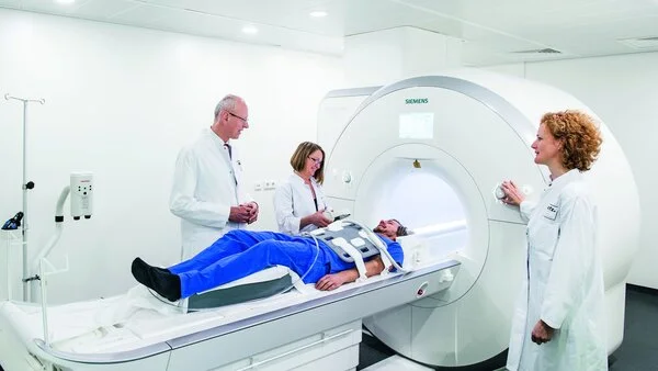 Kernspintomographie, © Nationales Centrum für Tumorerkrankungen (NCT); Foto: Tobias Schwerdt Ein Patient liegt auf der Liege eines Kernspinntomographen, während Ärzte neben ihm stehen und den Vorgang erklären.