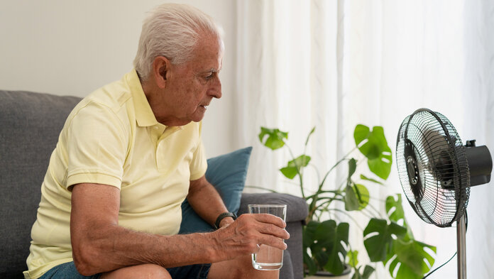 Ein älterer Mann sitzt auf dem Sofa vor einem Ventilator mit einem Glas Wasser.