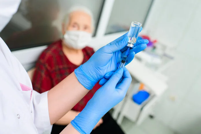 Man sieht die Hände einer Ärztin, die an einem Injektionsfläschchen eine Spritze aufzieht; im Hintergrund sitzt eine alte Patientin mit Mundschutz.