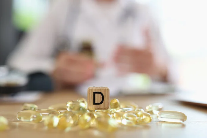 Auf einem Tisch liegen Kapseln mit dem Holzbuchstaben D, im Hintergrund verschwommen ein Arzt.