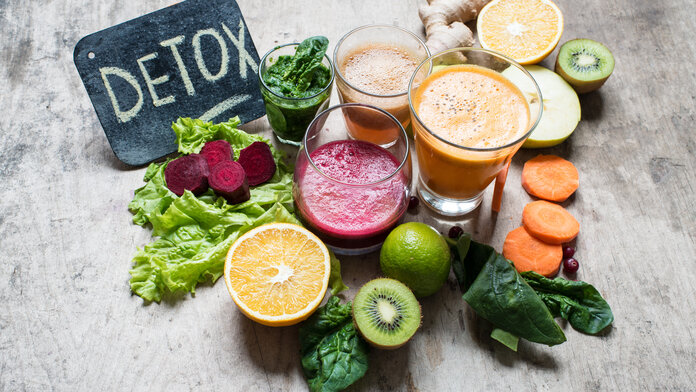 Auf einem Tisch steht ein Schild, auf dem “Detox“ geschrieben steht. Davor befinden sich Gläser mit verschiedenen Säften und geschnittenes Obst und Gemüse.