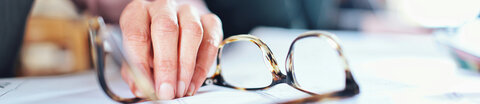 1© unsplash, Edan Cohen Eine Hand liegt neben einer Brille auf einem Schreibtisch. Im Hintergrund sieht man verschwommen andere Personen im Büro.