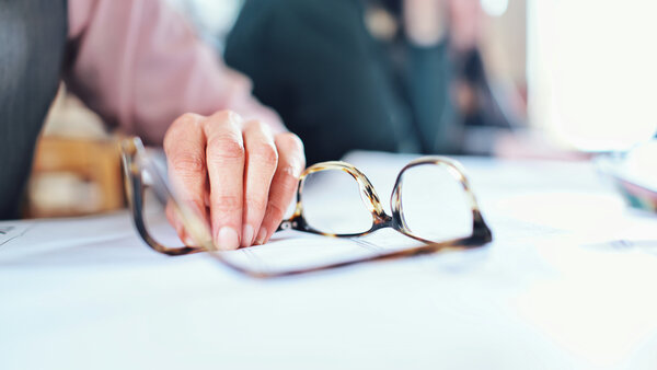 Eine Hand liegt neben einer Brille auf einem Schreibtisch. Im Hintergrund sieht man verschwommen andere Personen im Büro.