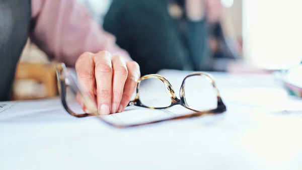 Eine Hand liegt neben einer Brille auf einem Schreibtisch. Im Hintergrund sieht man verschwommen andere Personen im Büro.
